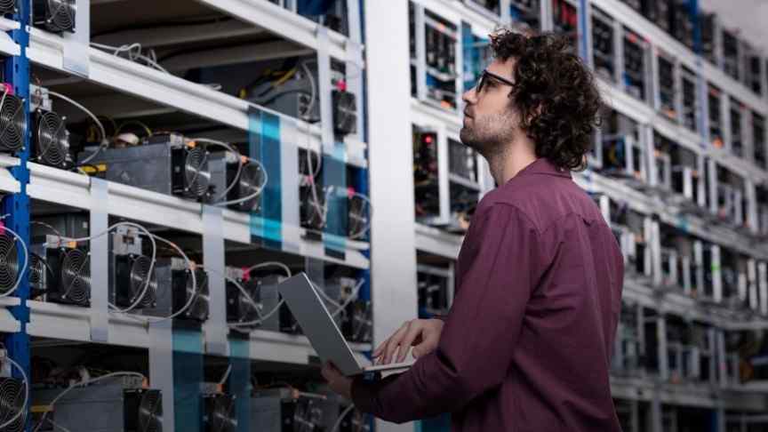 man in purple shirt and glasses holding laptop looking at mining rigs