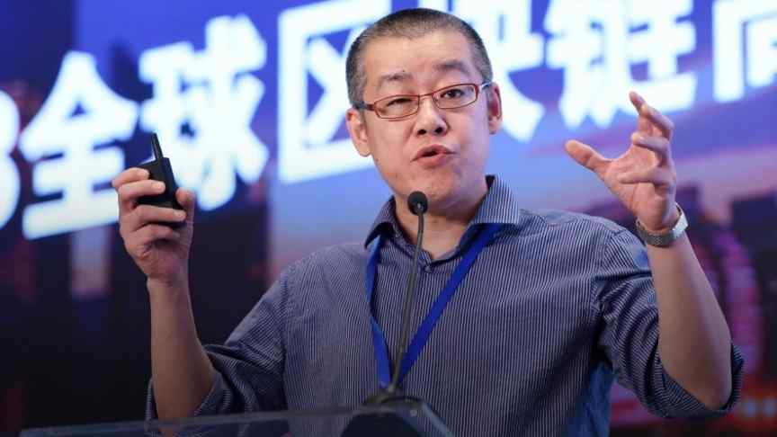 Li Xiaolai in grey shirt, raised hands, speaking into a microphone, chinese writing in the back