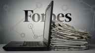 Forbes logo behind a desk with stacked newspapers and a laptop