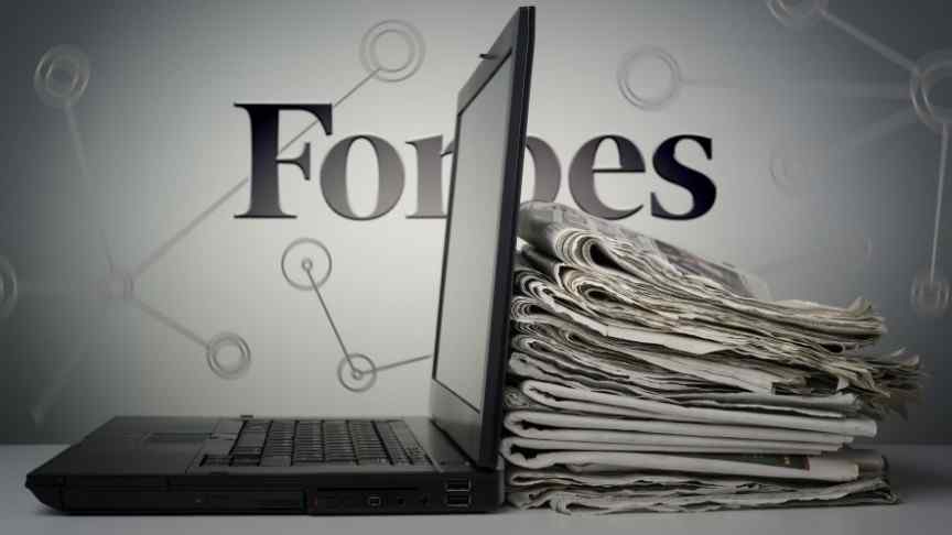 Forbes logo behind a desk with stacked newspapers and a laptop