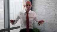 Nigel Green in white shirt, red tie, hands held up, sitting inside room with window and brick wall