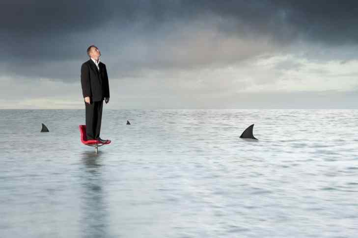 Suited man standing on a chair in the middle of the ocean, while sharks are near sighted