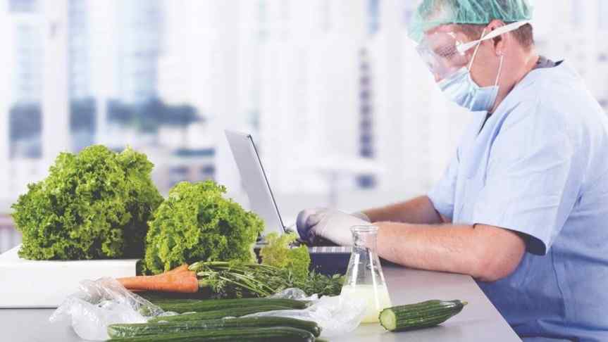 Doctor wearing a face mask and protection glasses sitting at a desk full of vegetables and operating a laptop