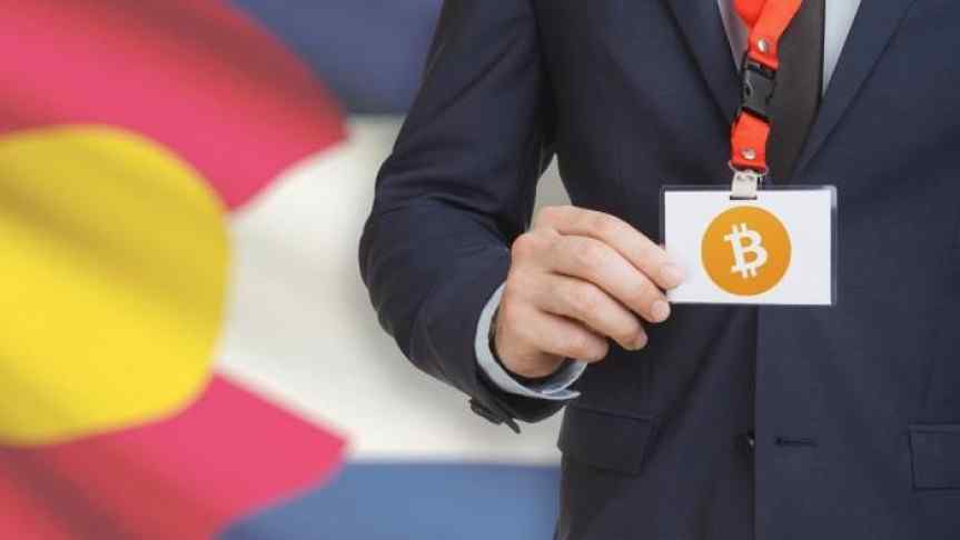 Suited man shoying his Bitcoin badge in front of the flag of the state Colorado