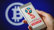 Hand holding smartphone showing FIFA 2018 Russia logo and PLACE A BET button on blue background with Bitcoin logo