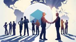 business people scattered on background of sunset and world map, two men at the front shaking hands. CryptoBrick logo in white circle.