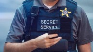 Man wearing grey shirt and Secret Service vest with radio and star logo, and holding phone in right hand.