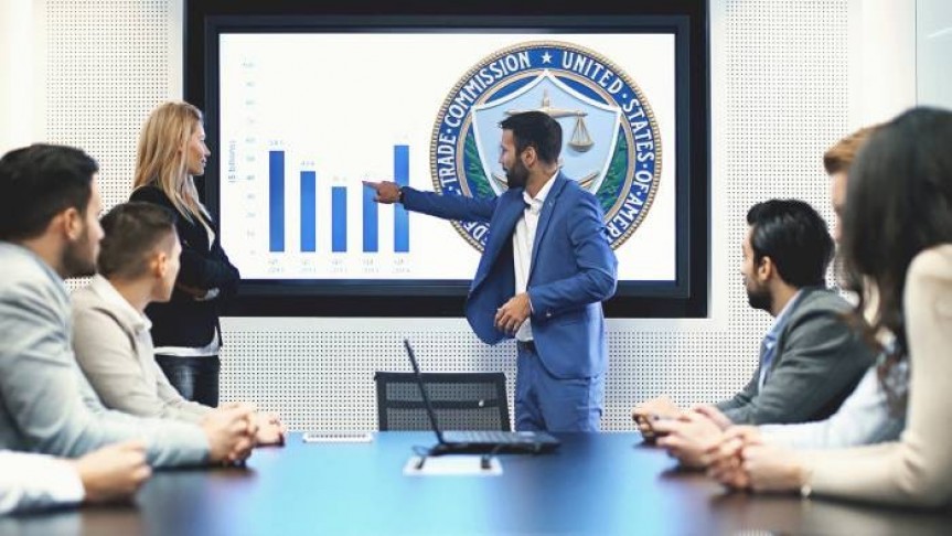 Man in blue suit pointing to graph and US FTC icon on screen in front of six people in suits sitting around office table.