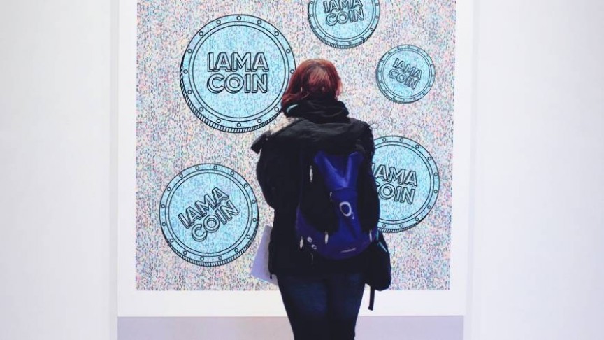 A woman wearing black and blue backpack standing in front of a painting of blue coins saying IAMACOIN on blurry background