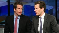 Winklevoss Twins sitting in an office. On the right in suit, blue shirt, red tie, black jacket. On the right, suit, white shirt, blue tie, grey jacket, mid speech