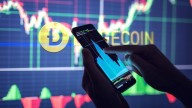 Dogecoin name and logo in yellow on background of screen showing graphs, hands holding smartphone also showing graphs