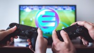 two sets of hands playing 2 PS4 controllers, TV screen showing football game and Enjin logo