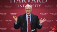 Oliver Hart in suit raising hands on red background of Harvard University wall